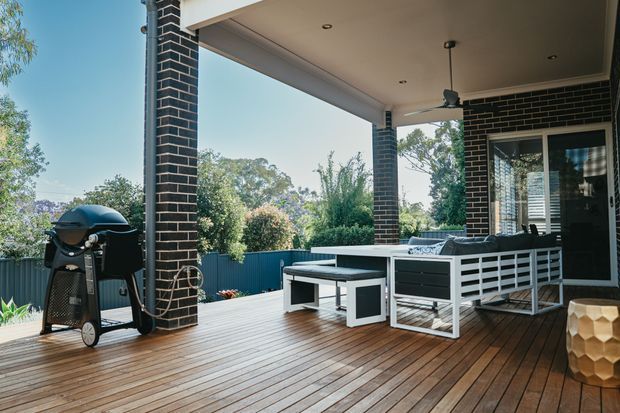 Covered outdoor entertaining deck with timber decking, dark brick columns and wall cladding, a ceiling fan, outdoor dining and lounge furniture, barbecue, and fenced garden surroundings. The space appears integrated with the house through large glazed doors.