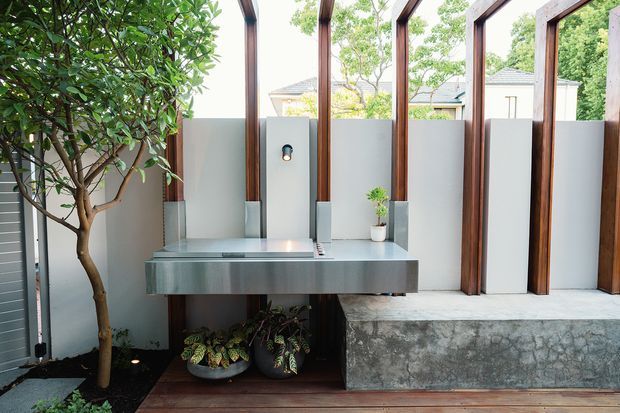 Contemporary outdoor kitchen and entertaining area with a stainless-steel counter and integrated cooktop, framed by tall timber posts and partially screened by pale grey privacy panels. A concrete bench, timber decking, potted plants, and a small tree create a refined indoor-outdoor setting.