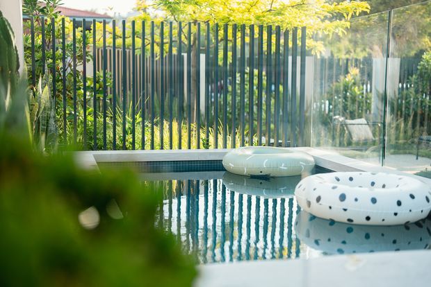 A sunlit outdoor swimming pool viewed close to the waterline, with blue-green tiled edging, reflective water, white inflatable pool floats, a transparent glass boundary, and a dark vertical-slat privacy fence surrounded by leafy greenery.