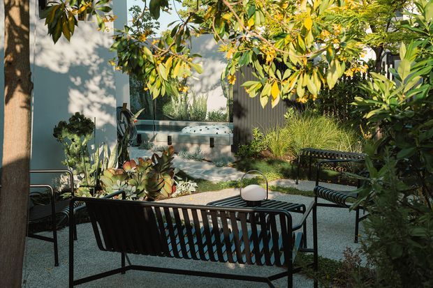 A leafy landscaped courtyard or outdoor lounge with black metal seating, a low table, gravel paving, abundant planting, and a small water feature or pool beside a contemporary building. Dappled sunlight creates a calm, private garden atmosphere.