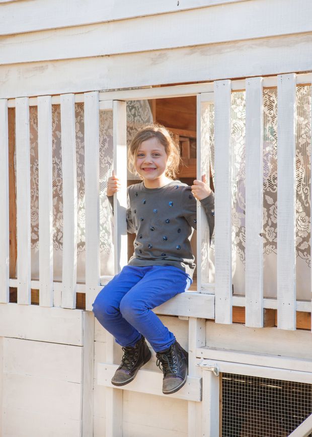 A bright exterior view of a small white-painted timber structure with horizontal weatherboard cladding and vertical slatted railing. Lace curtains and a timber-framed opening are visible behind the railing, while a child sits on the built-in sill or steps. The image focuses on the structure and its playful outdoor-facing enclosure.