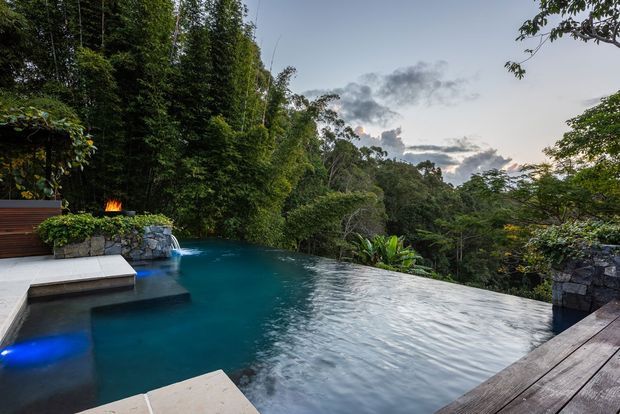 An infinity-edge swimming pool surrounded by dense tropical landscaping and mature trees, with dark timber decking, stone retaining walls, a small fire feature, and integrated blue pool lighting. The view looks out over a lush wooded setting beneath a cloudy evening sky.