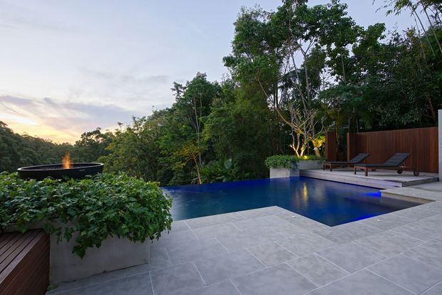 A landscaped outdoor pool terrace set within dense native greenery at dusk. The deep-blue swimming pool has a shallow stepped section, surrounded by large pale-grey tiled paving, timber decking, a dark timber privacy screen, sculptural planters, and reclining loungers. A raised fire bowl or water feature sits to the left, with warm landscape lighting among the trees.