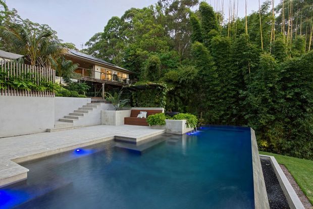 A landscaped residential outdoor area featuring a large modern swimming pool with illuminated blue underwater lights, broad pale paving, stepped access, raised planting beds, and a covered timber-clad house set among dense tropical greenery.
