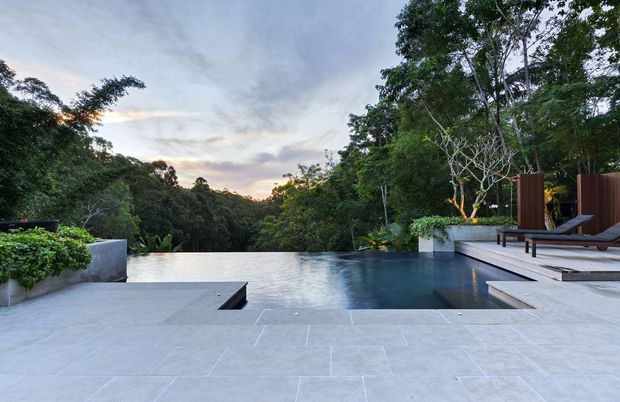 A luxurious outdoor infinity pool terrace overlooking dense tropical greenery at dusk. The broad pale stone-paved deck includes a dark reflecting pool, built-in raised planters, contemporary timber privacy screens, and lounge chairs. The scene has a calm, resort-like atmosphere and a strong connection to the surrounding landscape.