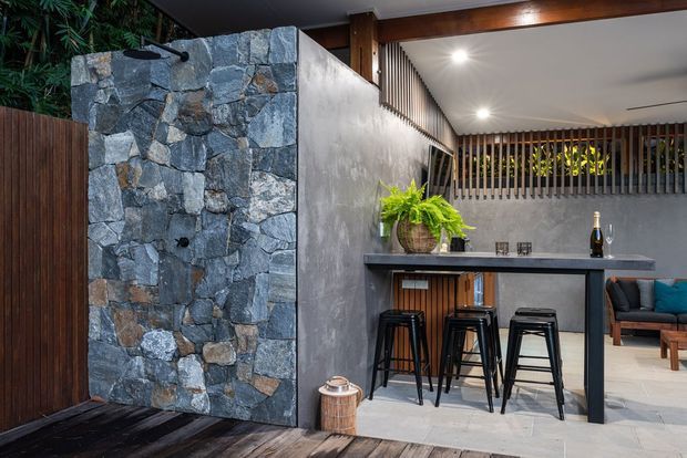 A sophisticated covered outdoor entertaining area featuring a rough natural-stone feature wall with water outlet, dark timber fencing and decking, a concrete-look bar counter with black stools, vertical timber screening, recessed lighting, and a lounge area beyond. The space has a refined resort-like atmosphere with integrated planting and warm timber accents.