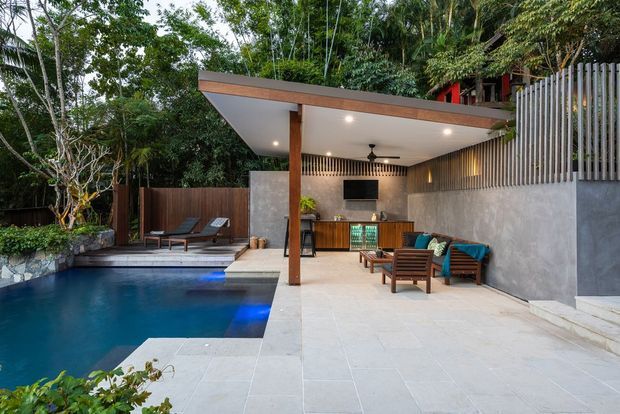 A contemporary covered outdoor entertaining area beside a swimming pool, set within dense tropical landscaping. The space features a flat timber-edged roof supported by a timber post, pale stone or concrete paving, a rendered masonry wall with vertical timber screening, a built-in outdoor bar or kitchenette, television, ceiling fans and recessed lighting. Timber lounge chairs and poolside seating create a relaxed resort-like setting.