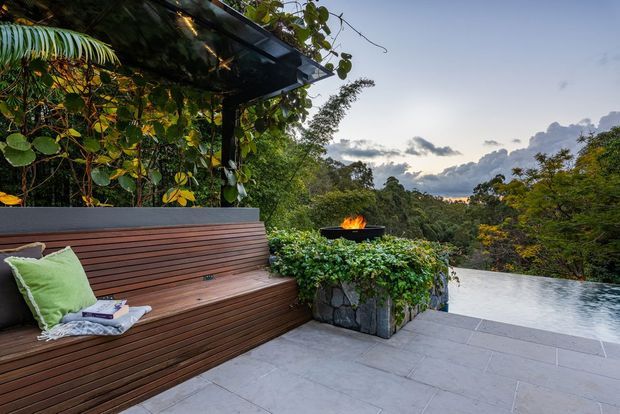 A landscaped outdoor terrace overlooking dense native bush, featuring a built-in timber bench beneath a leafy pergola, a raised stone planter with a fire bowl, and a pale tiled deck extending toward an infinity-edge pool or water feature. The setting is lush, private, and serene, photographed in soft evening light.