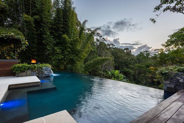 A secluded infinity-edge swimming pool surrounded by dense native vegetation, with dark timber decking, stone retaining walls, submerged blue lighting, and a small outdoor fire feature. The pool overlooks a lush forested landscape beneath a cloudy evening sky.