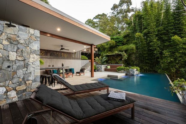 A contemporary outdoor entertaining area beside a swimming pool, set within dense greenery. The covered pavilion has a flat roof with timber detailing, exposed stone walls, timber posts, recessed lighting, a ceiling fan, an outdoor dining area and lounge seating. A dark timber deck surrounds the pool, with a small waterfall feature and stone planters.