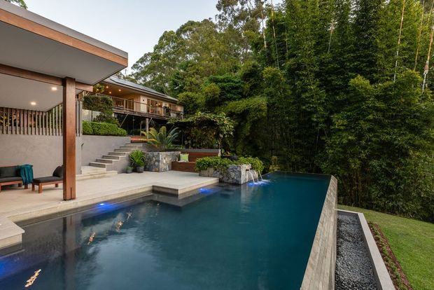 A landscaped outdoor pool area beside a contemporary multi-level home, featuring a dark tiled infinity-edge pool, timber and rendered architectural elements, stepped paving, stone planters, and dense bamboo and tree planting. The space has a refined resort-like feel and appears to be built on a sloped, elevated site.