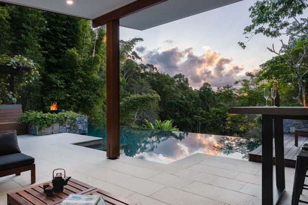 A covered outdoor entertaining terrace overlooking a lush forested landscape, with a reflective infinity-edge swimming pool, pale tiled paving, timber structural posts and beams, a built-in outdoor fireplace, lounge seating, and a high bar table. The scene is photographed at dusk with warm firelight and sunset clouds, creating a refined tropical retreat atmosphere.