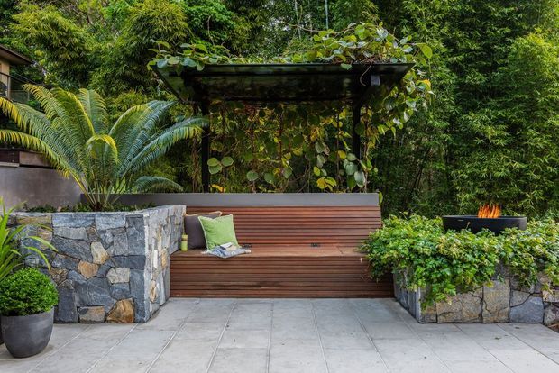 A landscaped outdoor courtyard or terrace featuring a built-in timber bench beneath a leafy pergola, surrounded by raised stone planters and lush tropical planting. A small fire feature is visible in a planter at right, and the ground is finished with large pale grey paving slabs.