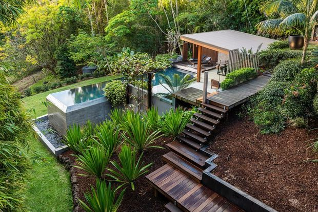 Elevated view of a modern pavilion-style outdoor living area set within dense tropical landscaping. The structure has a low-pitched flat roof, timber posts, extensive open glazing or screening, and a raised timber deck connected by stepped boardwalks. A compact rectangular swimming pool sits beside the pavilion, with planted garden beds, retaining walls, and lush vegetation surrounding the site.