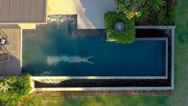 Aerial view of a long rectangular swimming pool beside a modern home, with dark pool edging, paved surrounds, landscaped garden beds, and a small circular outdoor feature set within dense greenery.