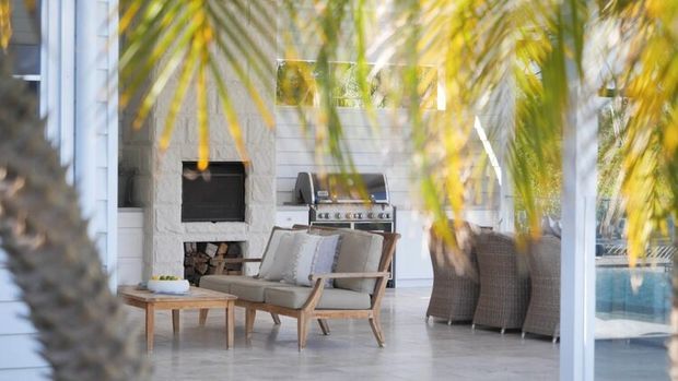 Bright covered outdoor living area with a white-painted masonry fireplace, built-in firewood storage, built-in stainless-steel barbecue, timber-framed lounge chairs, a coffee table, and wicker dining chairs. Palm foliage partially screens the space, and a swimming pool is visible beyond the glazing.