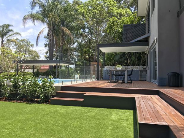 A contemporary residential outdoor entertaining area with a raised timber deck, built-in outdoor kitchen and bar seating beneath a covered pavilion. Steps lead down to a manicured lawn and a glass-fenced swimming pool, surrounded by lush tropical landscaping. The home has dark modern cladding and extensive outdoor living connections.