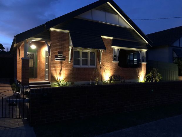 A dusk exterior view of a compact brick bungalow-style house. The facade features dark horizontal weatherboard cladding in the gable, a pitched gable roof with exposed eaves, brick walls, timber-framed windows, a recessed front porch, and warm accent lighting along the entry and garden bed. A low brick boundary wall and metal gate sit in the foreground.
