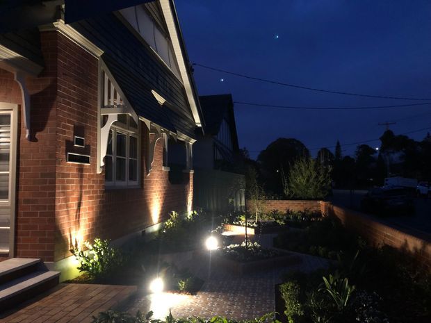 Twilight exterior view of a traditional brick home with decorative white trim, gabled roofline, and illuminated front garden. A brick-paved path and low landscaped planting lead toward the entrance, with neighbouring residential buildings and street infrastructure visible in the background.
