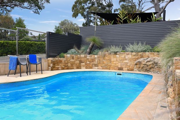 Outdoor residential swimming pool with bright blue water, surrounded by pale stone paving, tiered sandstone retaining walls, dark horizontal slat fencing, and drought-tolerant ornamental planting. A small outdoor seating area with colourful chairs sits beside the pool.