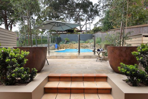 A landscaped residential courtyard and pool area viewed from a tiled patio. Broad terracotta-tiled steps lead past large rusted metal planters and clipped shrubs toward a small blue swimming pool, shaded by a large umbrella and enclosed by timber and corrugated metal fencing.