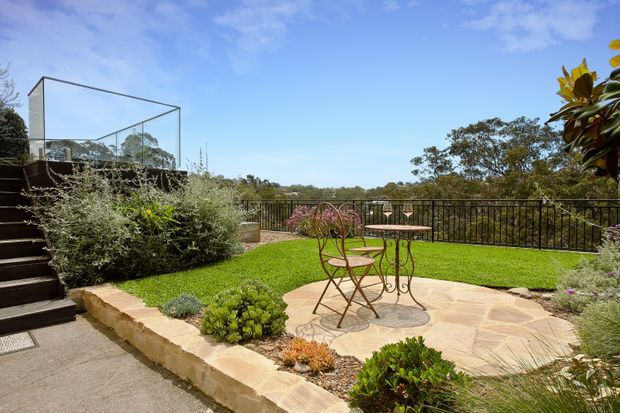 A landscaped residential outdoor terrace with a sandstone-paved seating area, ornate metal café table and chairs, manicured lawn, planted garden beds, and a black metal balustrade overlooking a wooded landscape. Steps lead up to a raised deck or terrace with transparent glass balustrading. The scene is bright and sunny with a relaxed garden atmosphere.