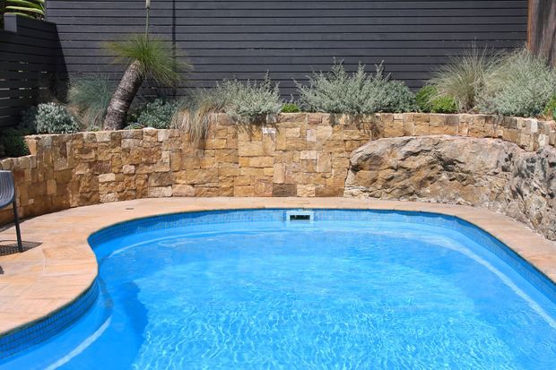 A bright outdoor swimming pool with curved edges, surrounded by terracotta-toned paving and a low natural stone retaining wall. The landscaped garden includes drought-tolerant grasses and succulents, with dark horizontal cladding forming the backdrop. A rocky outcrop is integrated into the poolside landscape.