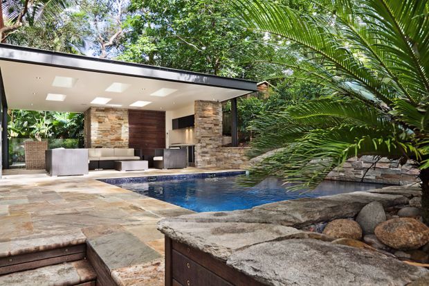A modern single-storey pavilion opens onto a landscaped pool terrace in a lush tropical setting. The covered outdoor living area includes built-in seating, a barbecue or outdoor kitchen zone, stone-clad columns and walls, and a flat roof with recessed lighting. Natural stone paving, boulders, dense planting and a deep blue swimming pool create a resort-like outdoor space.
