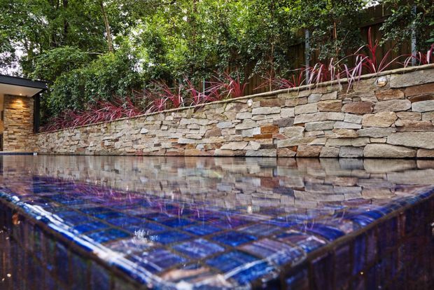A close-up view of a small outdoor pool or spa with deep blue tiled surfaces, reflecting the surrounding greenery and a rustic stacked-stone retaining wall. Dense planting and a timber privacy fence sit behind the wall, while a small stone-clad structure is visible at the left edge.
