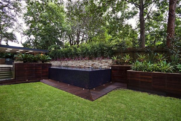 A landscaped backyard with a raised rectangular water feature or pool clad in dark textured material and topped with a natural stacked-stone edge. Timber decking borders the feature, with large dark timber planter boxes filled with tropical foliage. A manicured lawn, timber privacy fencing, dense mature trees, and a partially visible covered outdoor structure create a lush garden setting.