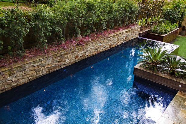 A landscaped outdoor swimming pool with vivid blue water, edged by stacked natural stone retaining walls and dark raised planter beds filled with tropical and ornamental plants. Dense hedging creates a private garden setting, while the pool includes integrated lighting and a narrow, contemporary layout.