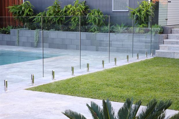 Modern residential outdoor area featuring a swimming pool, pale concrete paving, a neatly maintained lawn, frameless glass pool fencing with metal supports, and grey block retaining walls. Dense tropical planting lines the dark cladded facade, with concrete steps leading upward at right.