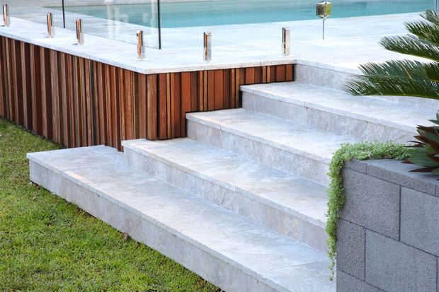 Exterior view of a landscaped pool area with broad pale stone steps leading to a raised swimming pool terrace. The pool is enclosed by clear glass balustrading with metal posts, while the terrace edge features vertical timber cladding and adjacent grey concrete blockwork. Tropical planting and lawn frame the stepped outdoor space.