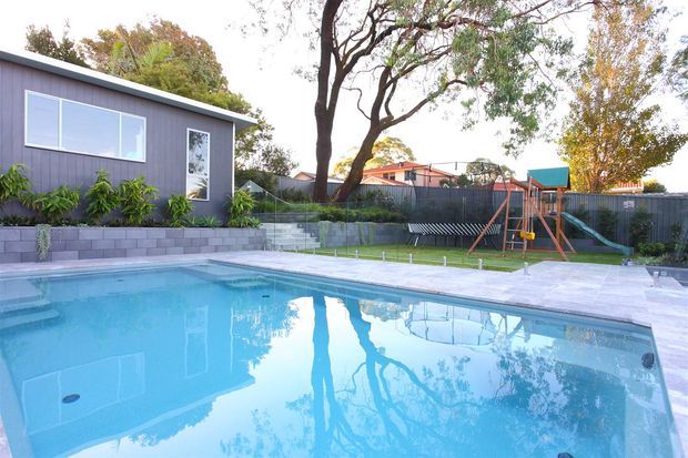 A suburban backyard swimming pool dominates the foreground, bordered by pale paving and a low grey retaining wall. Behind it is a contemporary dark-grey pool house or extension with large windows, landscaped planting, fencing, and a children's play structure. Mature trees and neighbouring residential buildings are visible in the background.