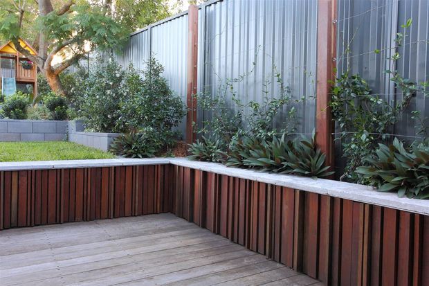 A landscaped residential outdoor courtyard or deck with built-in timber planter and bench edging. Vertical dark timber slats form the raised planter walls, paired with grey concrete block retaining walls and a tall corrugated metal privacy screen framed in timber. Dense planting softens the enclosed space, with a timber deck in the foreground.