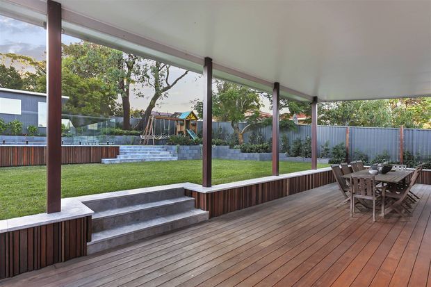A spacious covered outdoor terrace with warm timber decking, a timber dining table and chairs, and a long built-in raised planter or seating edge. Dark timber posts support a broad flat roof, while steps lead down to a manicured lawn and landscaped backyard enclosed by a metal fence.