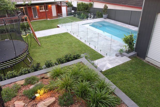 Elevated view of a landscaped residential backyard with a rectangular swimming pool, pale stone paving, frameless glass pool fencing, tiered garden beds, lawn, a trampoline, and a compact red outbuilding. The scene has a contemporary outdoor entertaining and recreational layout.