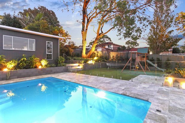A landscaped backyard with a large rectangular swimming pool in the foreground, surrounded by pale stone paving. The pool area is bordered by a low glass safety fence and overlooks a lawn with children's play equipment, garden planting, mature trees, and a dark-clad single-storey garden or poolside structure. Warm exterior lighting creates an inviting evening atmosphere.