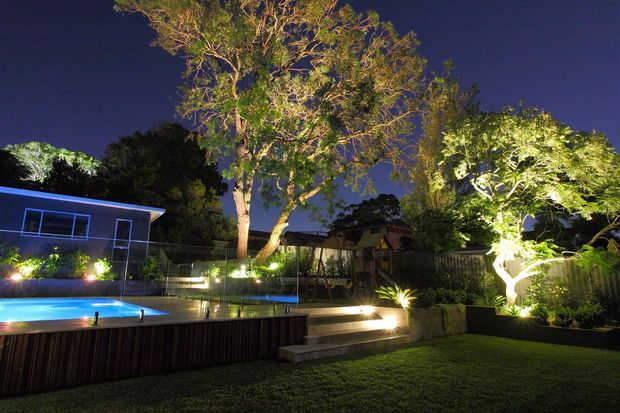 Night-time view of a landscaped residential backyard with a lit swimming pool, timber-clad pool surround, stepped deck access, illuminated mature trees, garden beds, fencing, and a low modern building visible at the rear.