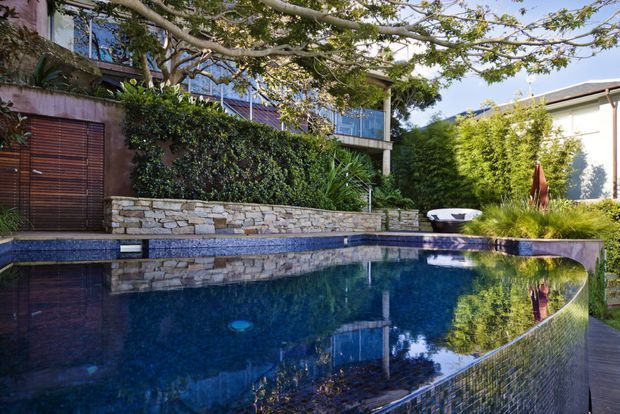 A landscaped residential courtyard dominated by a deep blue swimming pool with a curved edge and tiled finish. Stone retaining walls, dense planting, timber slatted gates, and a modern multi-level home with extensive glazing surround the pool, creating a private, leafy outdoor setting.