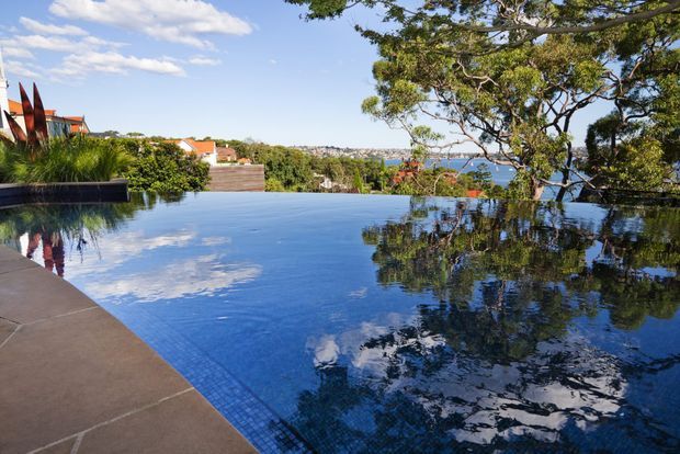 A tranquil infinity-edge swimming pool with dark blue tiled finish, reflecting the sky and surrounding trees. The pool overlooks a leafy suburban coastal setting with glimpses of water and distant buildings, while a paved terrace and landscaped planting frame the foreground.