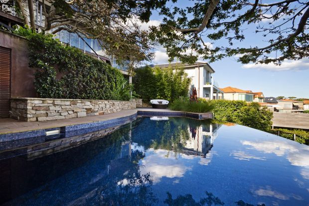 A landscaped residential swimming pool beside a modern multi-level home. The pool has dark blue tiled finishes and reflects the surrounding trees, sky and white building. A low natural-stone retaining wall, dense hedging, timber decking and mature trees frame the pool area.