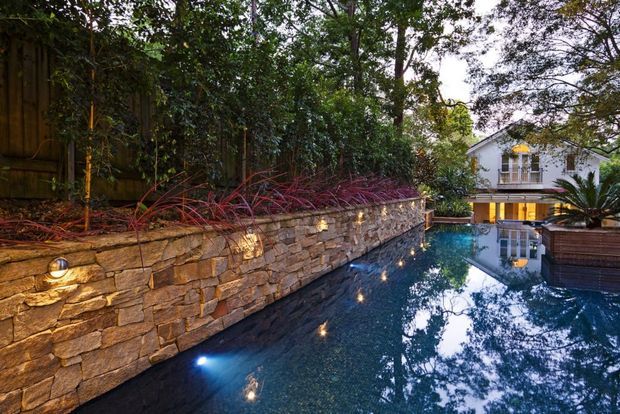 A landscaped residential swimming pool beside a tall natural-stone retaining wall with integrated warm lighting. Dense planting lines the wall, while a contemporary timber-clad pool structure and a light-coloured multi-storey house are visible beyond the water. The pool reflects the surrounding trees and architecture in a calm twilight setting.