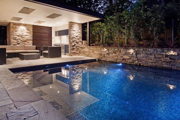 Twilight view of a contemporary outdoor entertaining area with a blue-tiled swimming pool, shallow lounging shelf, and integrated stone paving. A covered open-plan lounge and bar area sits beside the pool, framed by stacked natural-stone walls and timber screening, with warm recessed ceiling lights and planted borders.