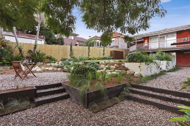 Landscaped residential courtyard with gravel ground cover, tiered dark timber garden beds, stone-filled planting areas, grasses and drought-tolerant plants. Stepped paths connect the different garden levels, with outdoor timber seating and a timber privacy fence. Brick and weatherboard residential buildings are visible around the courtyard.