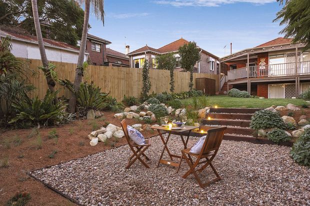 A landscaped residential courtyard or backyard with a compact gravel seating area furnished with a small wooden folding table and two chairs. Stone-edged steps with subtle lighting lead through layered planting toward surrounding homes, timber fencing, and brick buildings with terracotta tiled roofs. The scene has a relaxed, warm outdoor entertaining atmosphere.
