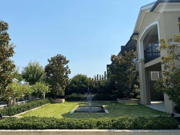 A landscaped courtyard garden beside a substantial multi-storey residential building. The formal lawn is bordered by neatly clipped hedges and mature trees, with a small rectangular reflecting pool and central fountain as the focal point. The building has a light rendered facade, tall arched openings, dark metal balcony railings, and a projecting roofline under a clear blue sky.