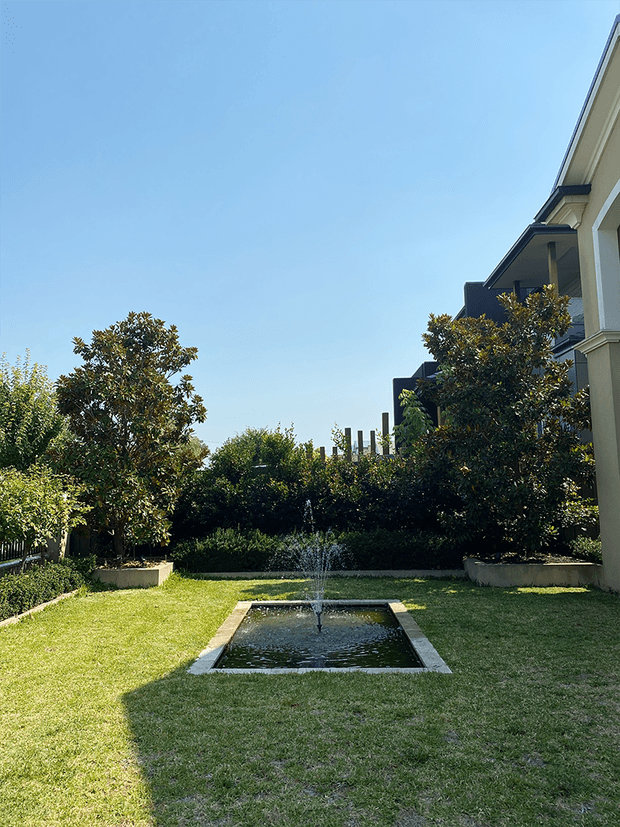 A formal landscaped courtyard garden beside a large residential building, featuring a rectangular reflecting pond with a central fountain, manicured lawn, clipped hedges, mature ornamental trees in raised planters, and a clear blue sky. The view also shows parts of the building facade and projecting balconies along the right side.