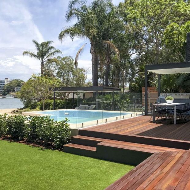 A landscaped waterfront outdoor living area with a clear glass-fenced swimming pool, broad timber decking, stepped platforms, and a covered dining pavilion. Tropical palms and mature planting surround the space, with water and nearby buildings visible beyond.