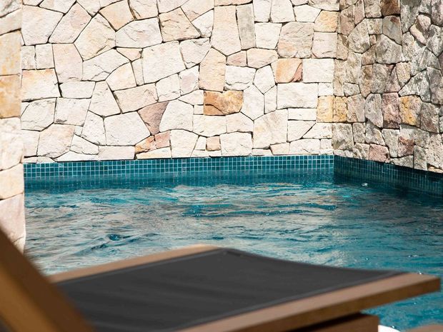 Close-up of a small swimming pool or spa with turquoise mosaic-tile edging and irregular pale natural-stone walls. A dark wooden poolside platform or lounge surface occupies the foreground. The scene has a warm, rustic Mediterranean character with strong turquoise and stone tones.